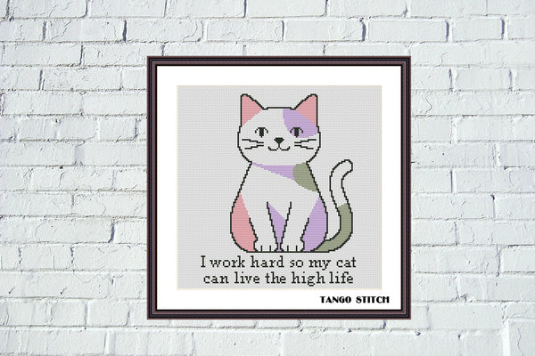 A framed cross-stitch style cat print hanging on a white brick wall. The artwork shows a stylized cat with pink, purple, white, and gray sections above the text, 'I work hard so my cat can live the high life. Humorous Pet Quote Embroidery cross stitch pattern - Tango Stitch