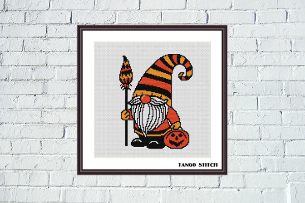Framed Halloween-themed cross-stitch pattern of a gnome with a striped hat and pumpkin on a white brick wall. Halloween gnome festive spooky cross stitch pattern - Tango Stitch